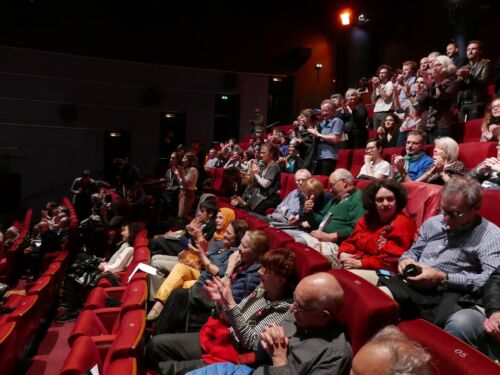 An audience sitting on red seats in a theatre.