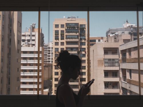 A silhouetted woman standing in front of a large window looking at her phone. Through the window you can see high rise housing blocks.