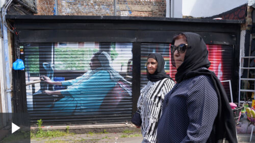 Two women pass in front of a mural on shop shutters