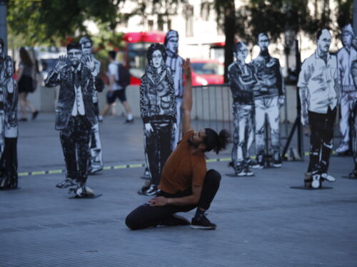 A male dancer wearing a brown short sleeved top and black trousers, kneeling with one arm stretched into the air. He is outside and surrounded by large cutouts of drawings of people.