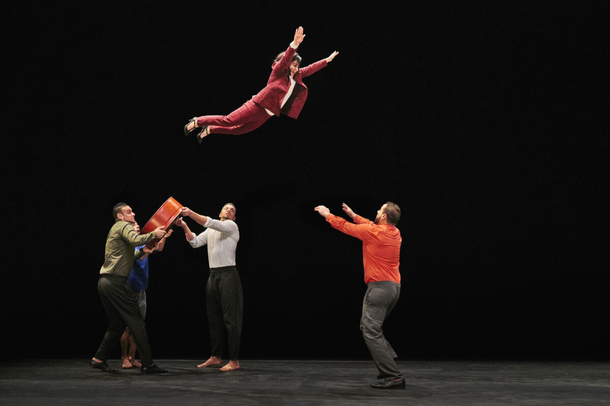 An image of a troupe of acrobats dressed in colourful everyday clothing. A woman is flying through the air and the catcher has his arms outstretched.