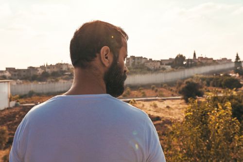 A man with brown hair and a beard wearing a white t-shirt with his back to the camera looking at the dividing wall in Jerusalem
