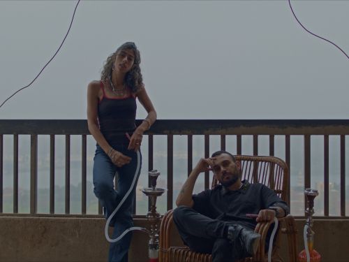 A portrait of a man and woman on a balcony. She is standing he is seated, both have shisha pipes.