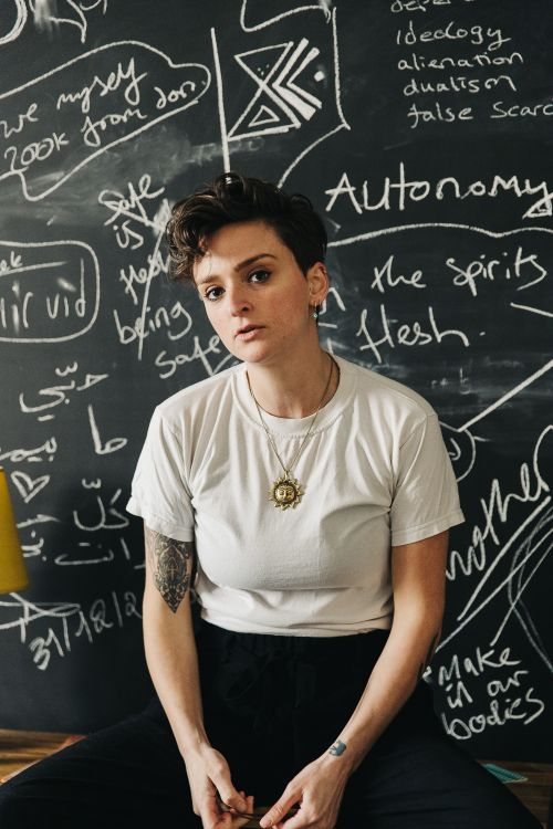 A woman with short dark hair wearing a white t-shirt sat in front of a blackboard covered in chalk writing