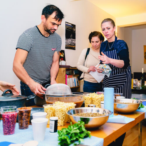 Three people standing in front of a table of ingredients including chickpeas, pasta, herbs and pickles