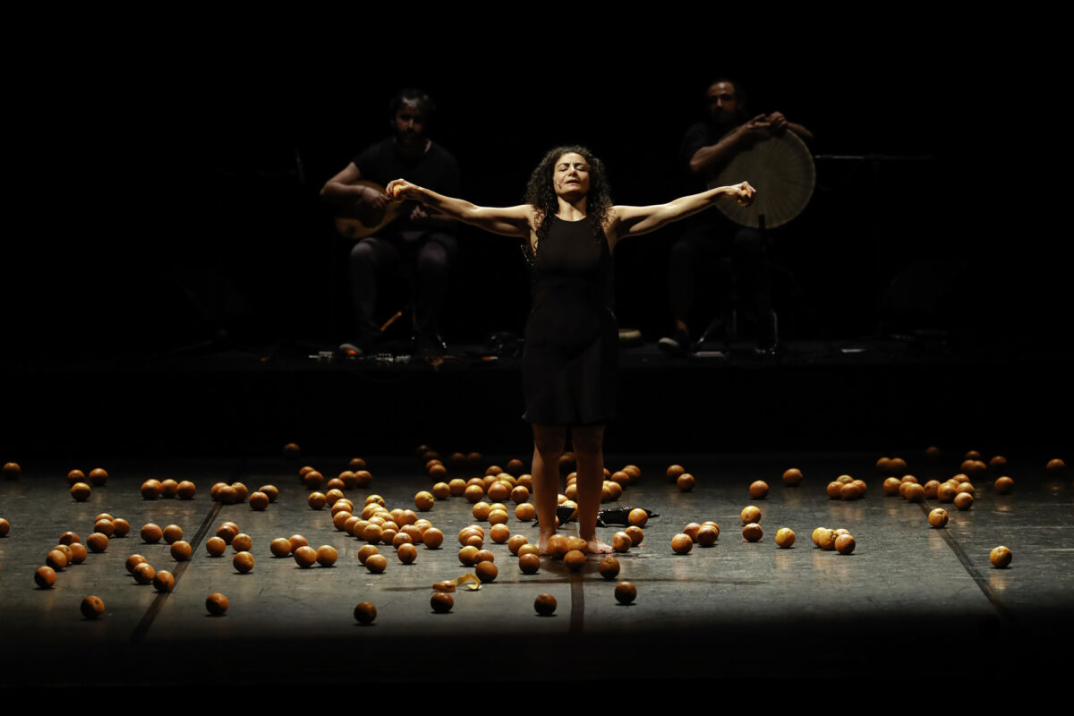 Image of woman standing in darkness on stage with her arms out wide with an orange in each hand. Many oranges strewn across the floor.