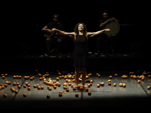 Image of woman standing in darkness on stage with her arms out wide with an orange in each hand. Many oranges strewn across the floor.