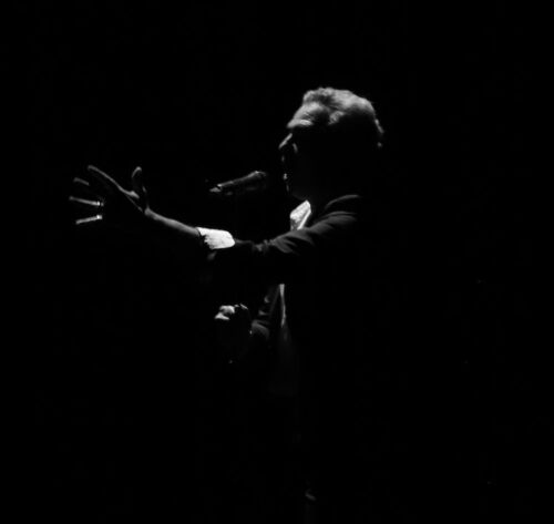 Man on stage reaching out into darkness