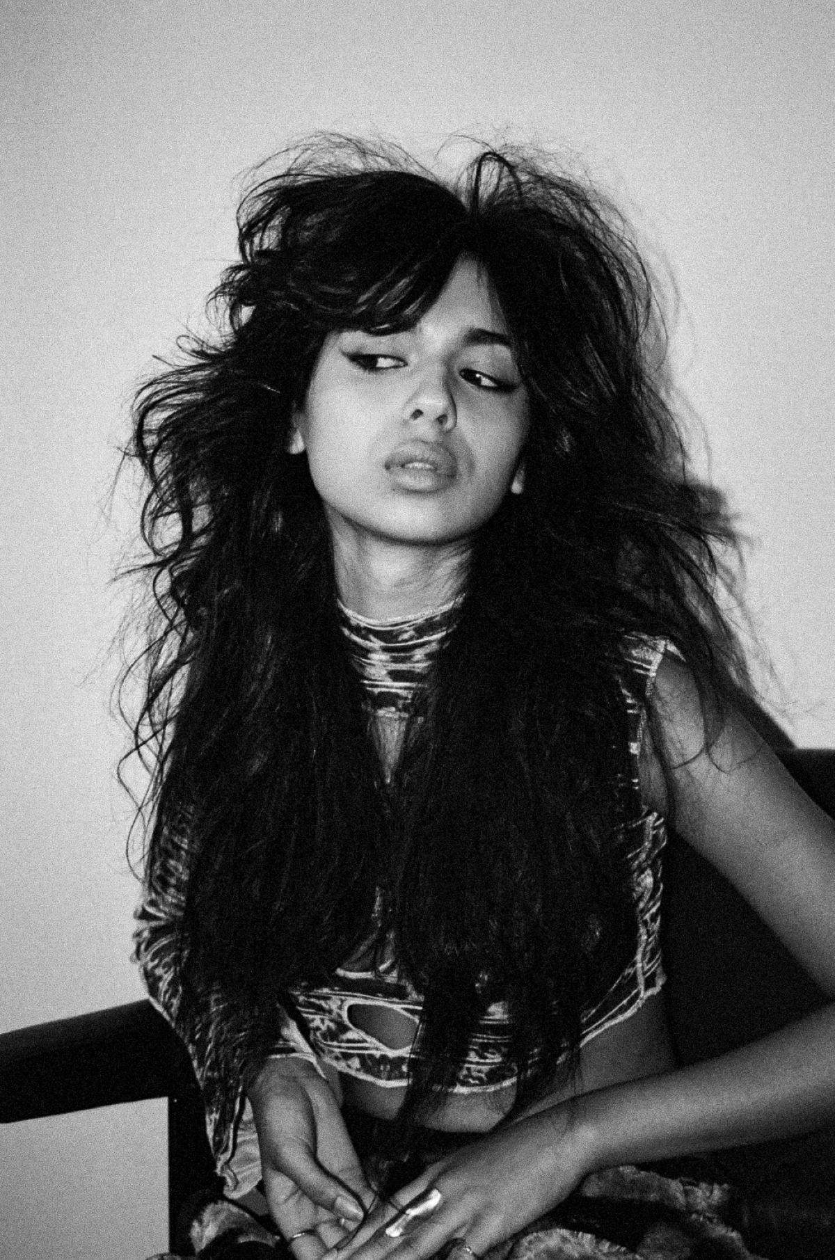 Black and white photo of a person with voluminous hair, wearing a patterned top, sitting with a contemplative expression, looking to the side. Vintage vibe.