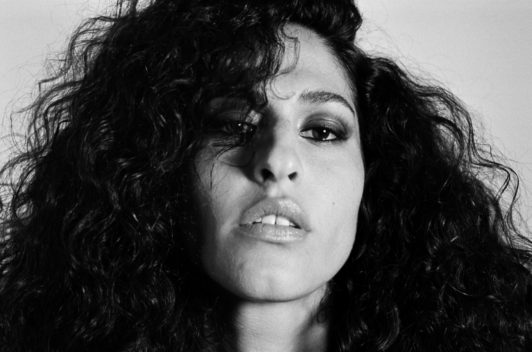 Black and white close-up portrait of a person with bold, curly hair and an intense gaze. The expression conveys confidence and strength.