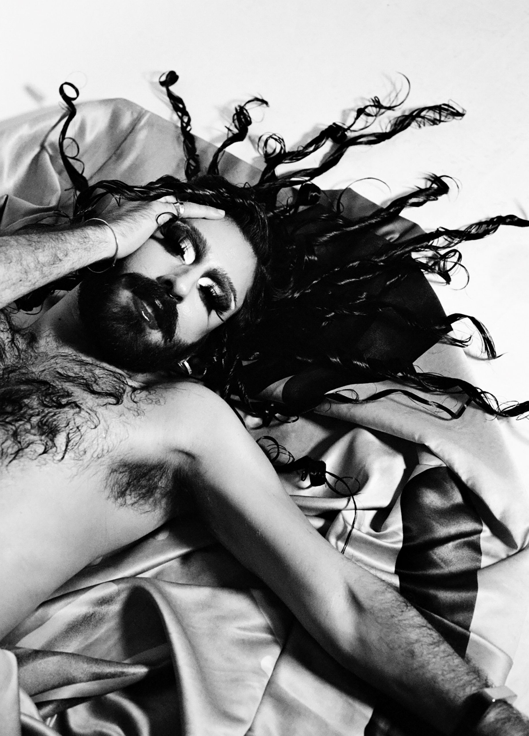 Black and white image of a person with dramatic makeup and curled hair, lying on satin sheets. They appear relaxed and contemplative.