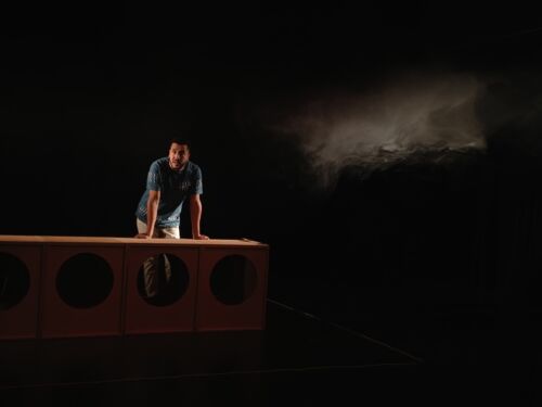 Man leaning forward with both hands on a minimalist stage set, looking ahead under soft theatrical lighting. A dark stage and drifting smoke create a dramatic atmosphere.