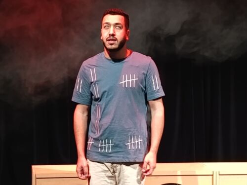 Man standing on stage with a focused expression beneath red and white theatrical lighting. Smoke fills the dark background behind a minimalist stage set.