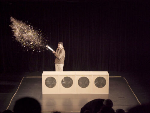 Man standing behind a minimalist stage set as a burst of golden confetti fills the air.